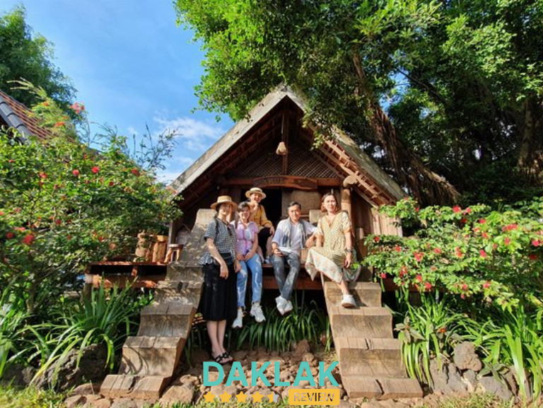 “Nhóm du khách chụp ảnh trước căn nhà dài Ê Đê trong khuôn viên Arul Coffee Buôn Ma Thuột, bao quanh bởi cây xanh và hoa rực rỡ.”