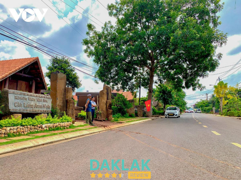 “Cổng vào buôn Akô Dhông ở Buôn Ma Thuột với bảng đá khắc tên buôn, nhà dài truyền thống và hàng cây lớn bên con đường yên bình.”