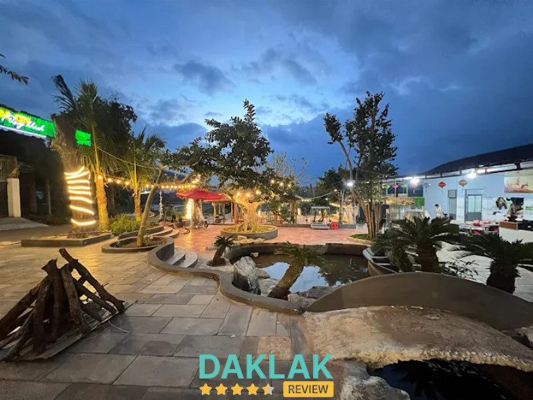 “Không gian Quán Nhậu Bến Nước Ea Kar buổi chiều tối, sân vườn rộng, trang trí đèn vàng và hồ nước tạo không khí thoáng mát.”